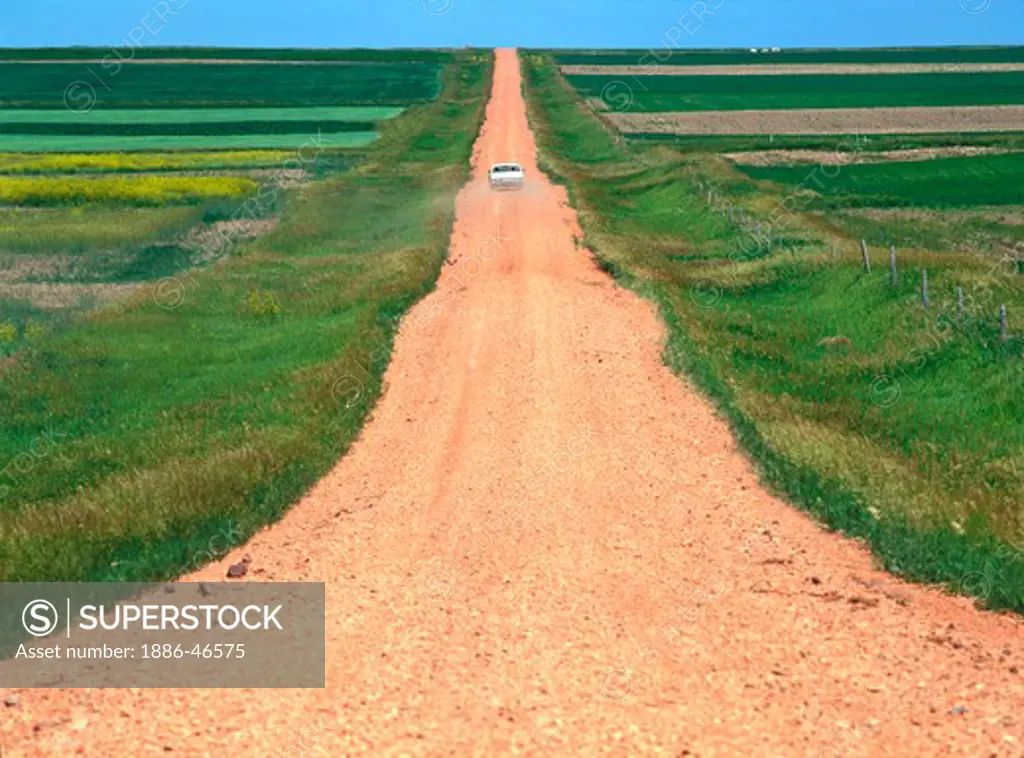 White sedan automobile travels a rural red crushed rock gravel road through farm fields in North Dakota, USA.
