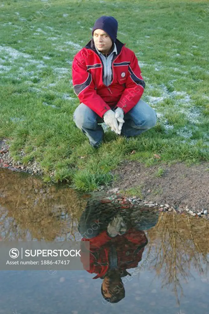 Young man sitting near pond with reflection in water snow on the grass Sweden, MR # 468