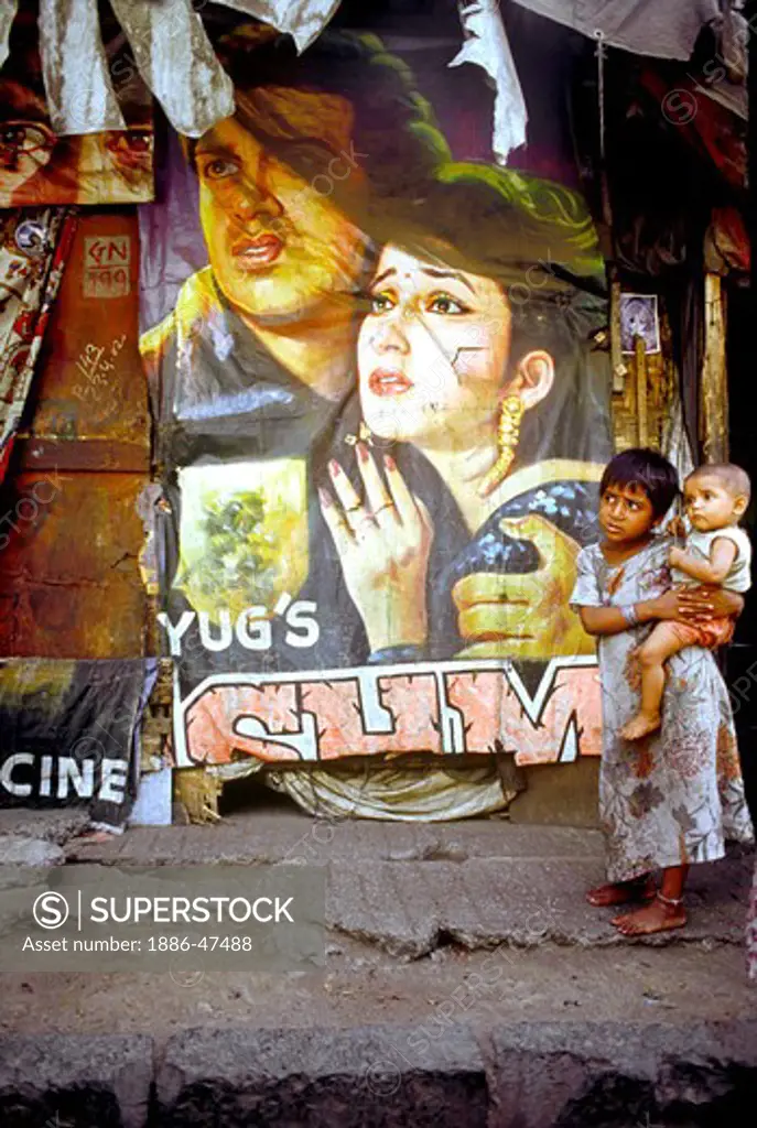 A young girl with her sibling in arm standing in front of her house and bollywood film poster ; Mumbai Bombay ; Maharashtra ; India