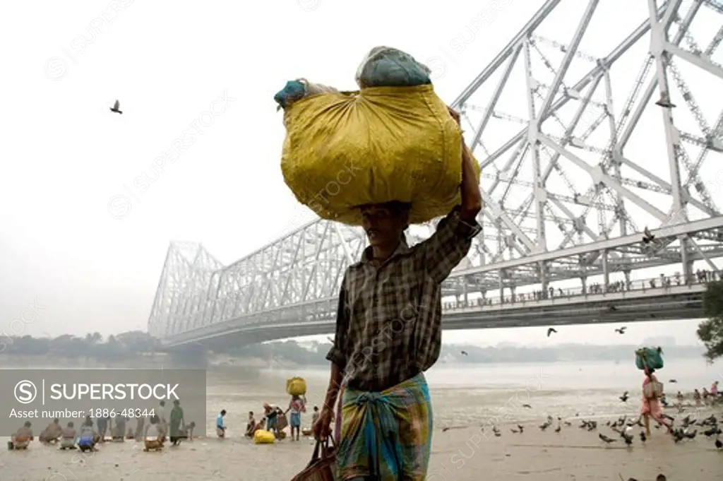Man carrying load on head Howrah bridge over Hooghly river in background ; Calcutta now Kolkata ; West Bengal ; India