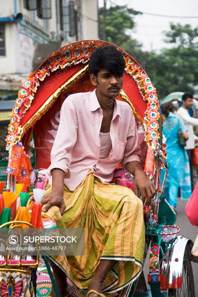 Cycle rickshaw  puller wearing lungi and shirt sitting on his vehicle waiting for passenger ; Dhaka ; Bangladesh