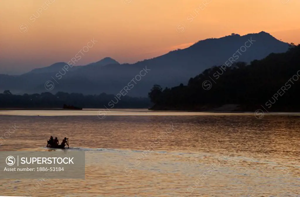 The sun sets over a hill on the Mekong River silhouetting a Laotian paddling their river boat as it runs through  - LUANG PROBANG,  LAOS