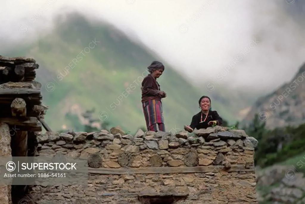 Two WOMEN wearing Dolpo Blankets unique to the area stand on the ROOF of a stone house in RINGMO village - DOLPO, NEPAL