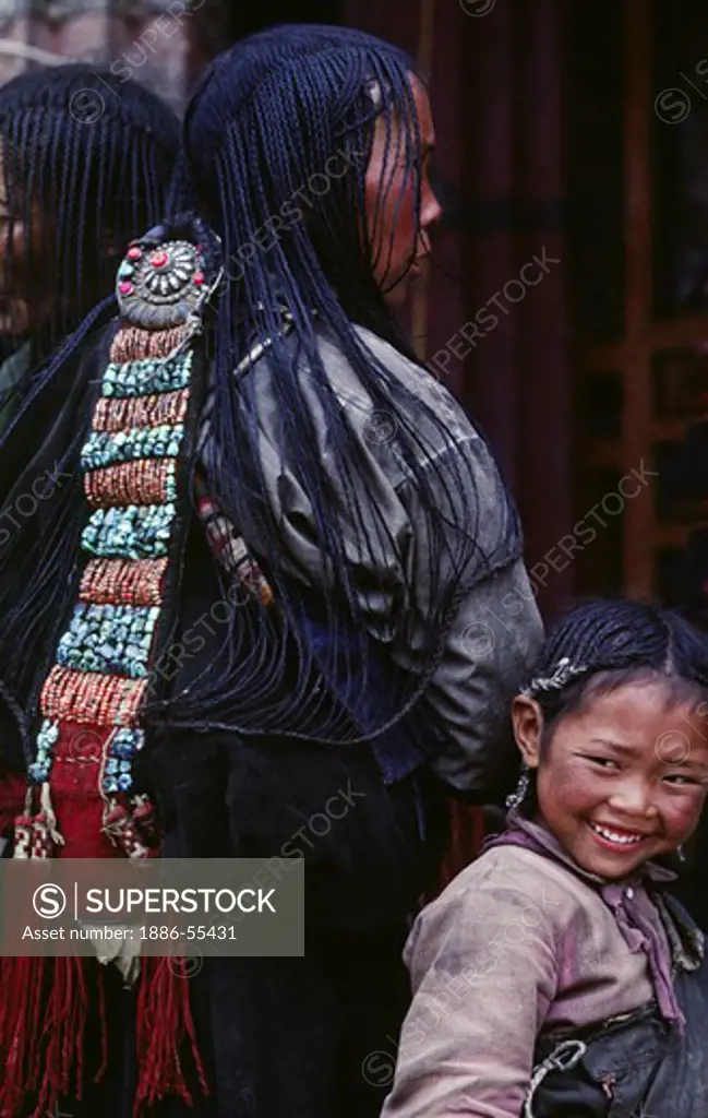 Young girl stands behind her mothers 108 braids & beautiful coral & turquoise hair piece - Sera Monastery, Lhasa, Tibet