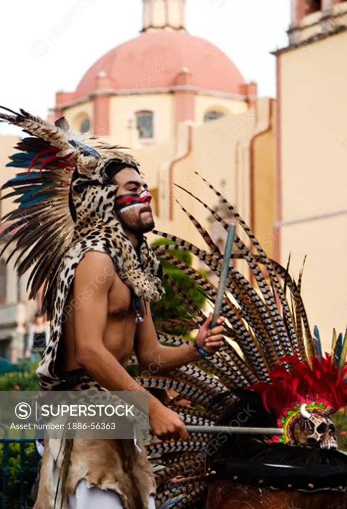 An AZTEC DANCER plays a DRUM dressed in a traditional warrior feathered COSTUME during the CERVANTINO FESTIVAL  - GUANAJUATO, MEXICO