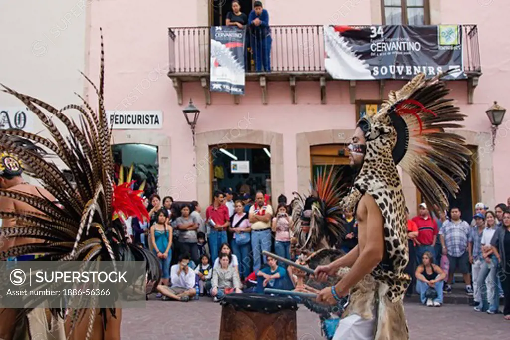 An AZTEC DANCER plays a DRUM dressed in a traditional warrior feathered COSTUME during the CERVANTINO FESTIVAL  - GUANAJUATO, MEXICO