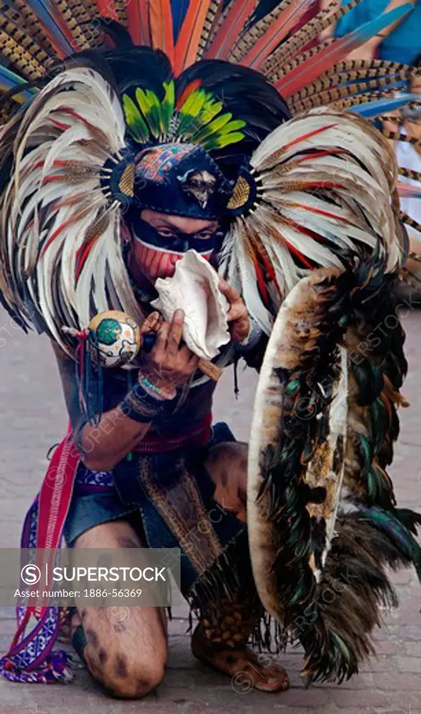 An AZTEC DANCER blows a CONCH in a traditional feathered warrior COSTUME during the CERVANTINO FESTIVAL - GUANAJUATO, MEXICO