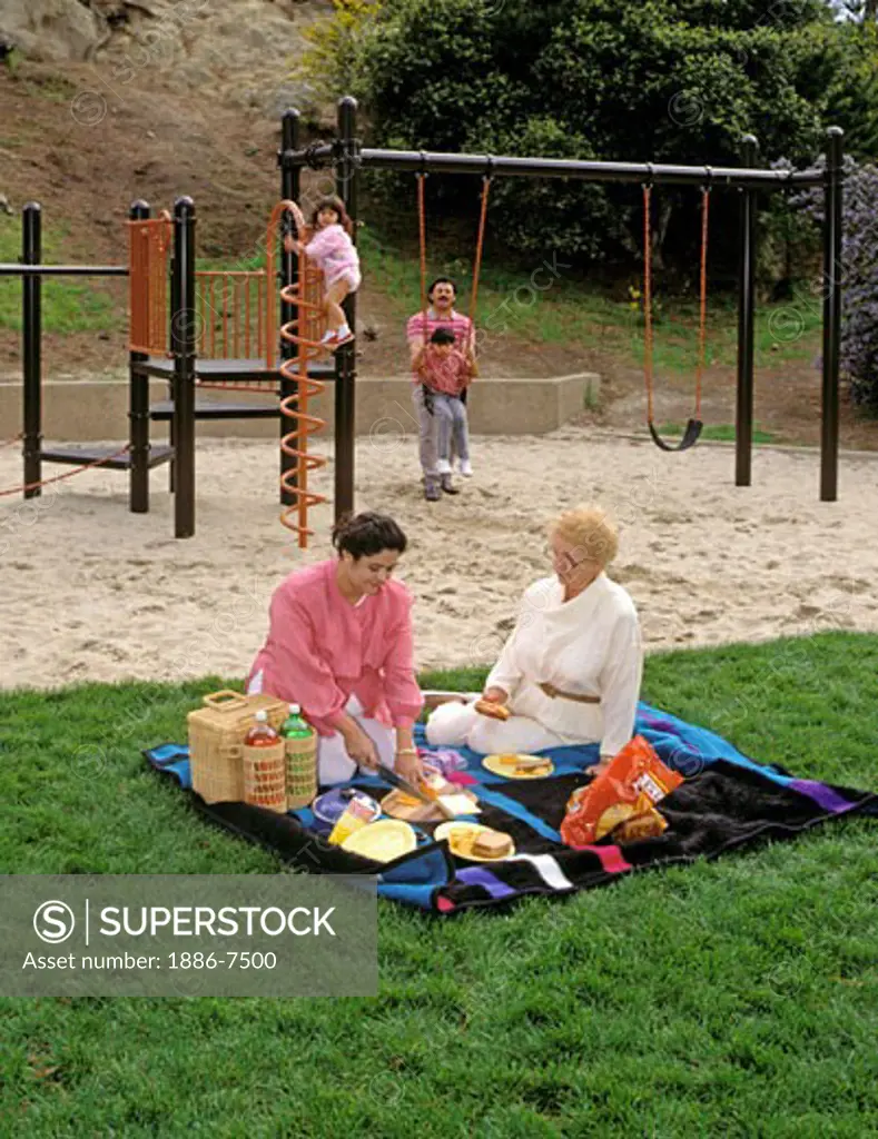 HISPANIC mother & grandmother set up PICNIC while father & children play on the PLAYGROUND 