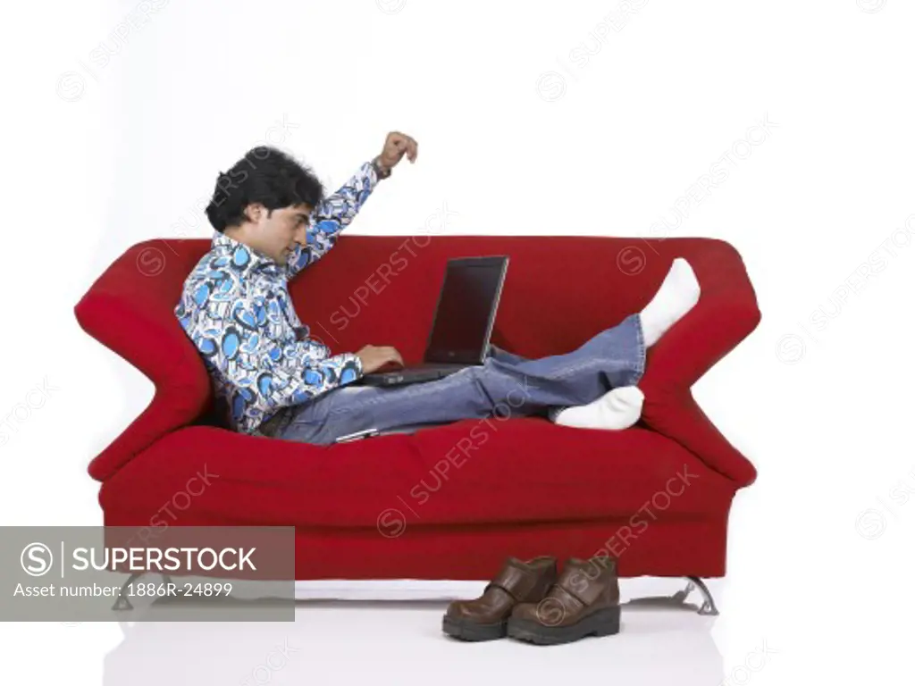 VDA200162 : South Asian Indian man sitting on sofa working on laptop wearing jeans and shirt, MR # 701