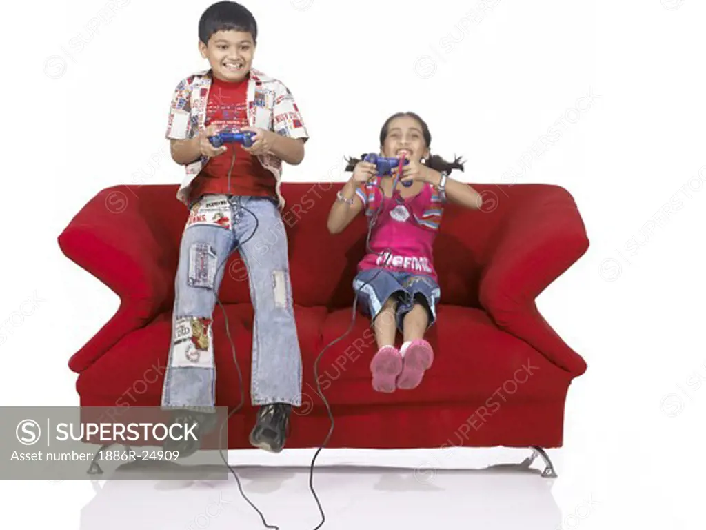 VDA200176 : South Asian Indian two children brother and sister jumping on sofa playing video game wearing jeans and shirt, MR #  699, 700