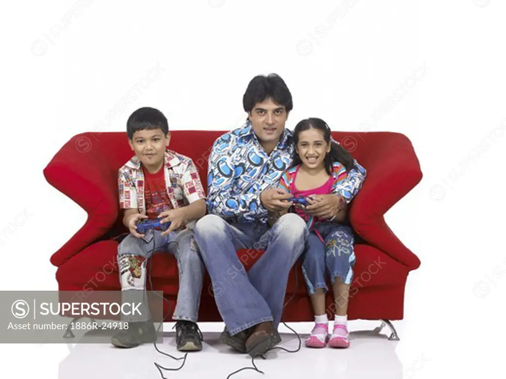 VDA200186 : South Asian Indian father with son and daughter sitting on sofa smiling, playing video game, enjoying and looking at camera wearing blue shirt and jeans, dress, MR #  699, 700, 701