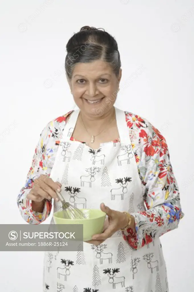 Senior Citizen Old woman holding light green coloured bowl and egg beater or hand mixer, smiling, wearing trousers, top with full sleeves and red, blue, yellow gray flowers and leaves, chain with pendent, earrings wearing apron with the images of reindeer, MR # 703A