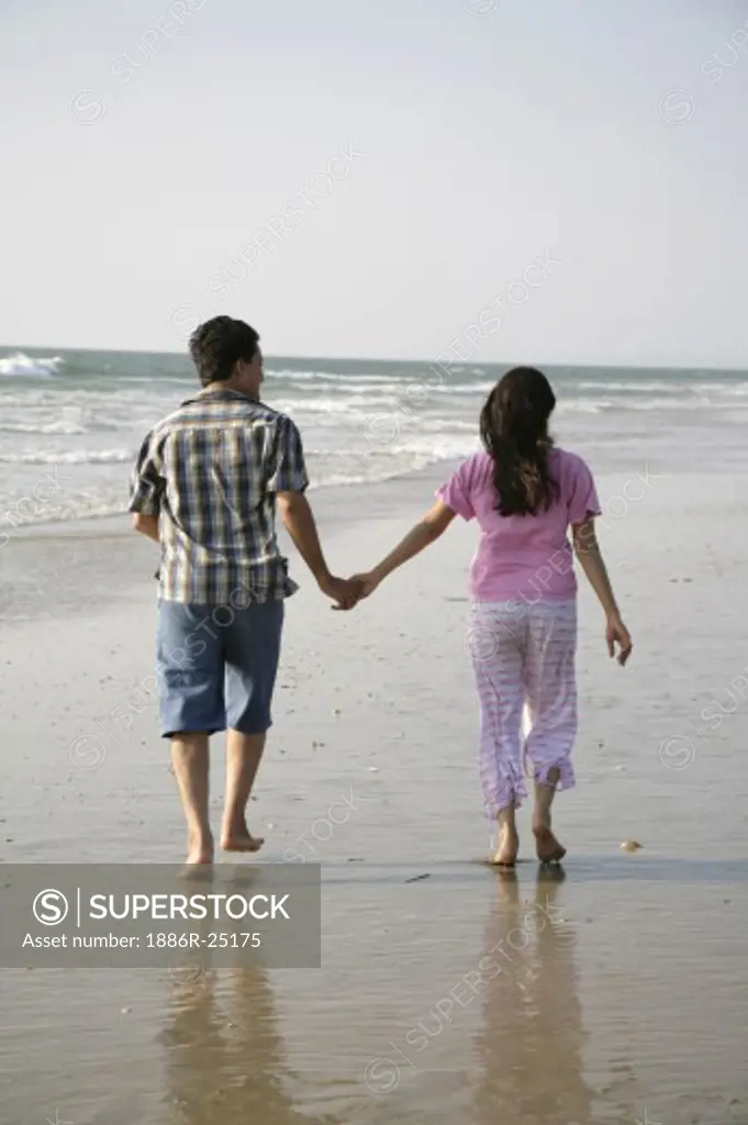 South Asian Indian young man and woman holding hands walking together on seashore; Shiroda; Dist. Sindhudurga; Maharashtra; India MR#703D; 703E