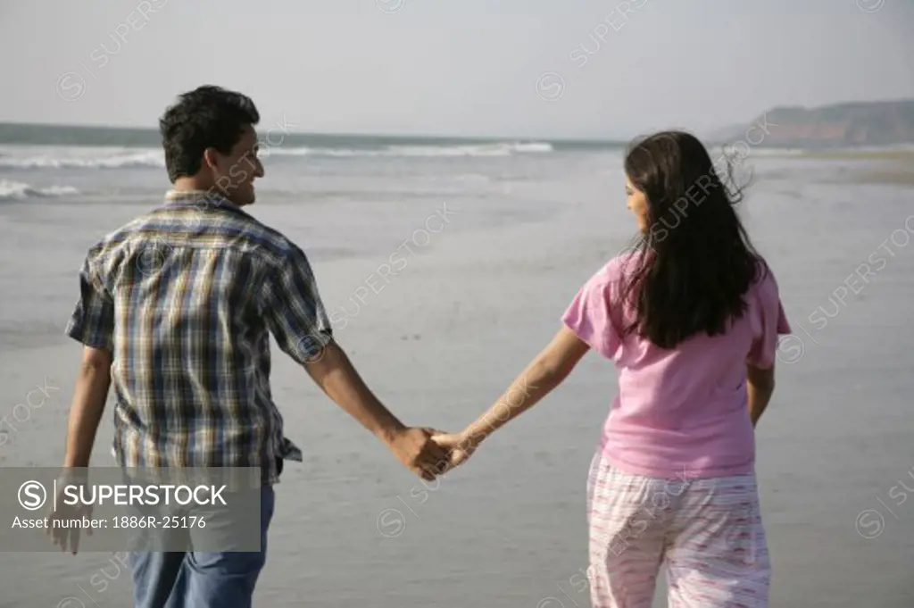 South Asian Indian young man and woman holding hands walking together on seashore; Shiroda; Dist. Sindhudurga; Maharashtra; India MR#703D; 703E