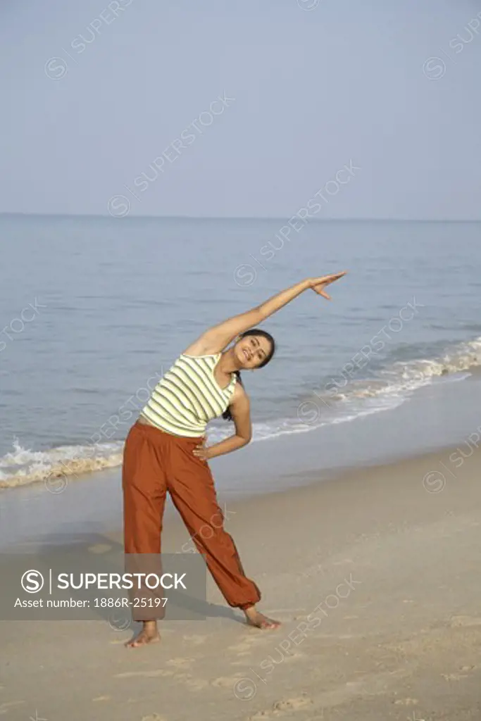 South Asian Indian young lady doing aerobics standing with raising one hand bend left side on seashore ; Shiroda ;  Dist. Sindhudurga ; Maharashtra ; India MR#703E