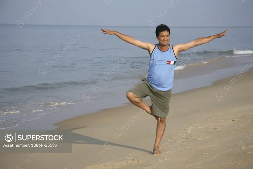 South Asian Indian young man doing aerobics standing on one folded leg with spread hands both side on seashore ; Shiroda ;  Dist. Sindhudurga ; Maharashtra ; India MR#703D