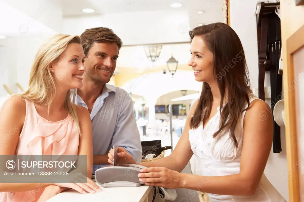 Female Sales Assistant At Checkout Of Clothing Store With Customers