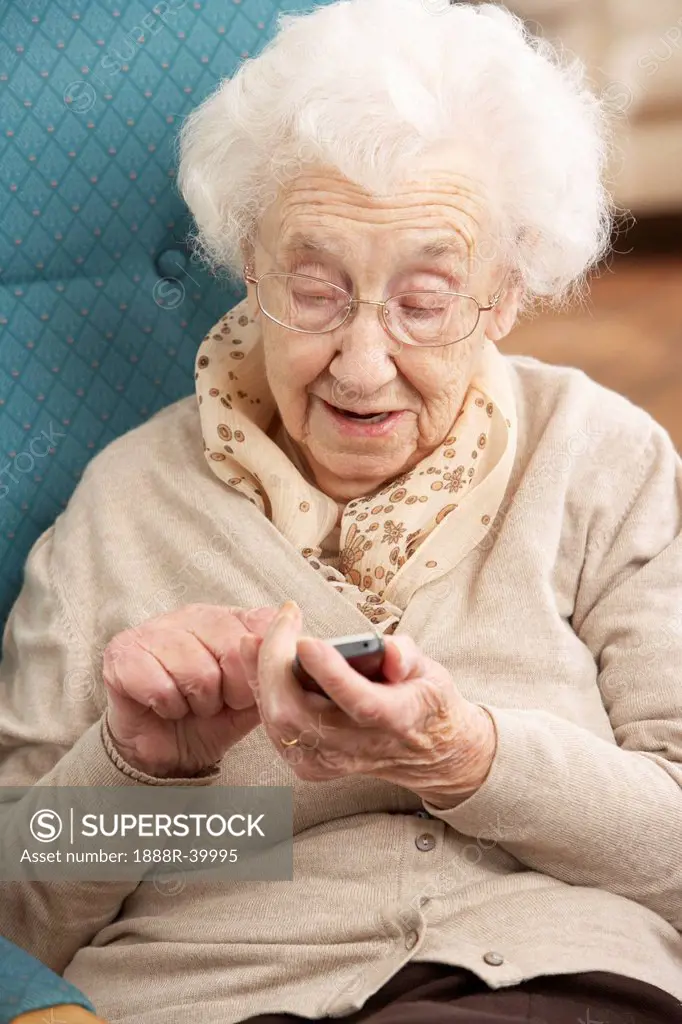 Senior Woman Dialling Number On Mobile Phone Sitting In Chair At Home