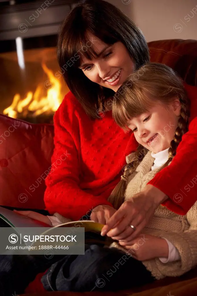 Mother And Daughter Sitting On Sofa And Reading Book By Cosy Log Fire
