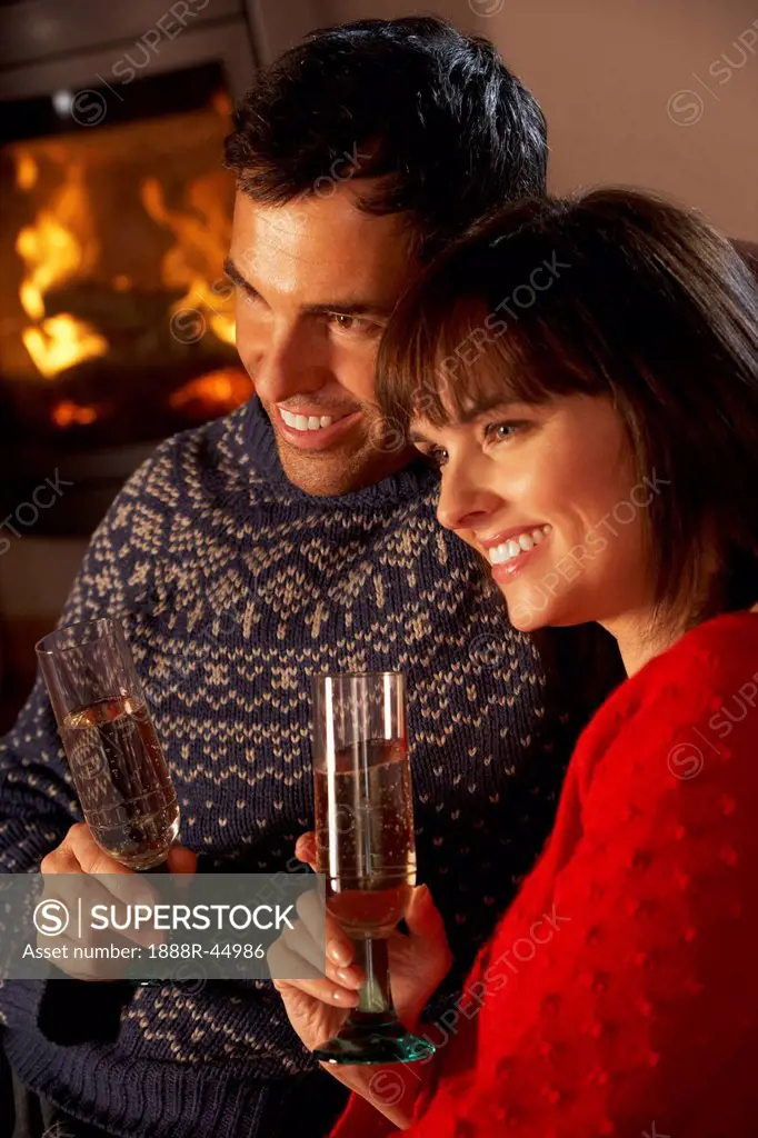 Middle Aged Couple Sitting Sofa By Cosy Log Fire With Glass Of Champagne