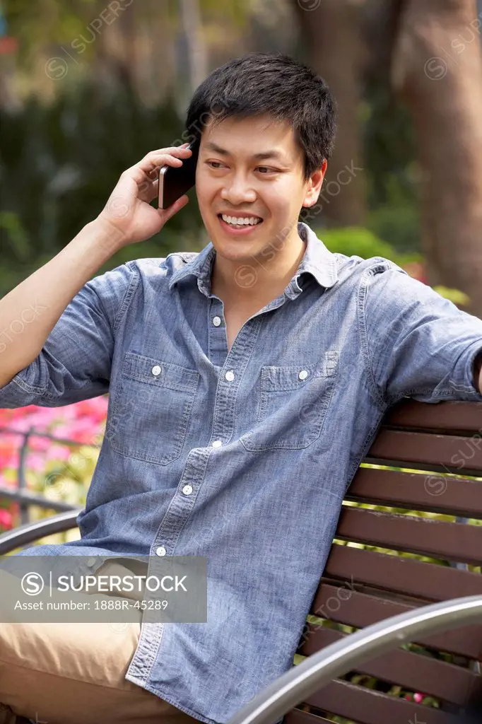 Young Chinese Man Relaxing On Park Bench Talking On Mobile Phone