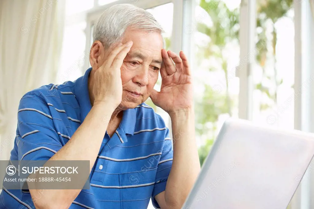 Worried Senior Chinese Man Sitting At Desk Using Laptop At Home