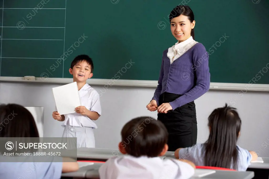 Pupil And Teacher Standing By Blackboard In Chinese School Classroom