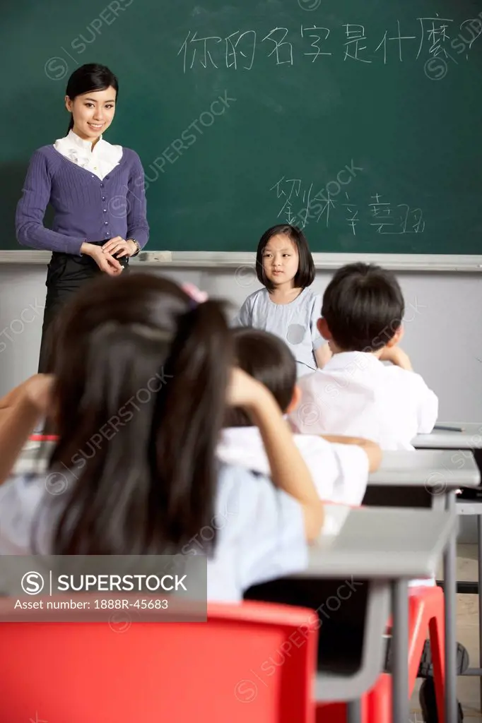 Pupil And Teacher Standing By Blackboard In Chinese School Classroom