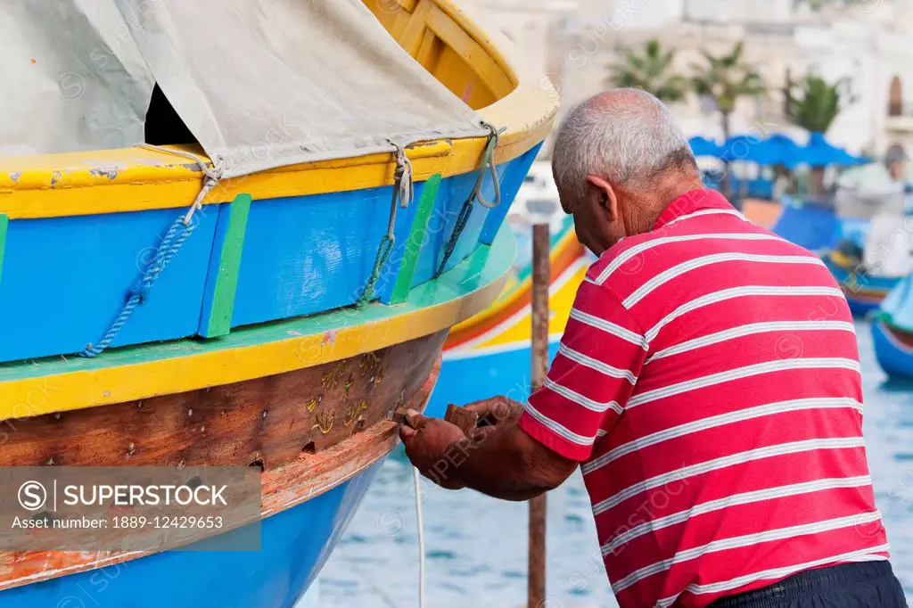 Fisherman Fixing His Luzzu, A Traditional Maltese Fishing Boat On The Beach, Marsaxlokk, Malta