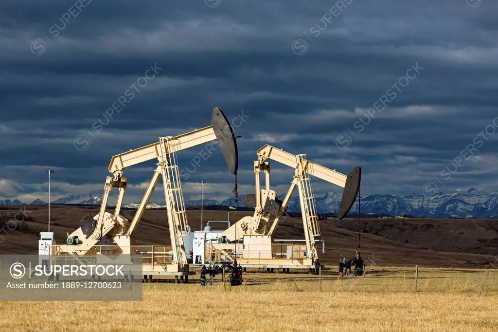 Two dramatically sun lit pumpjacks in a field with dark clouds in the sky and shadowed mountains in the background; Alberta, Canada