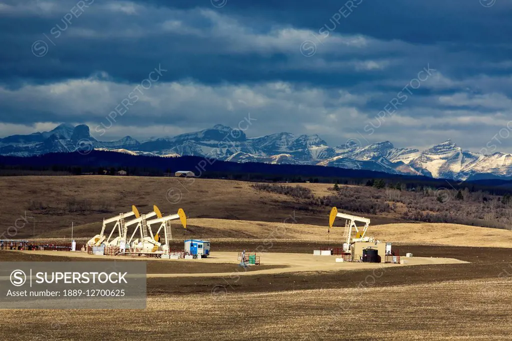 Sun lit pumpjacks in a field with dark clouds in the sky, shadowed rolling hills and mountains in the background; Alberta, Canada