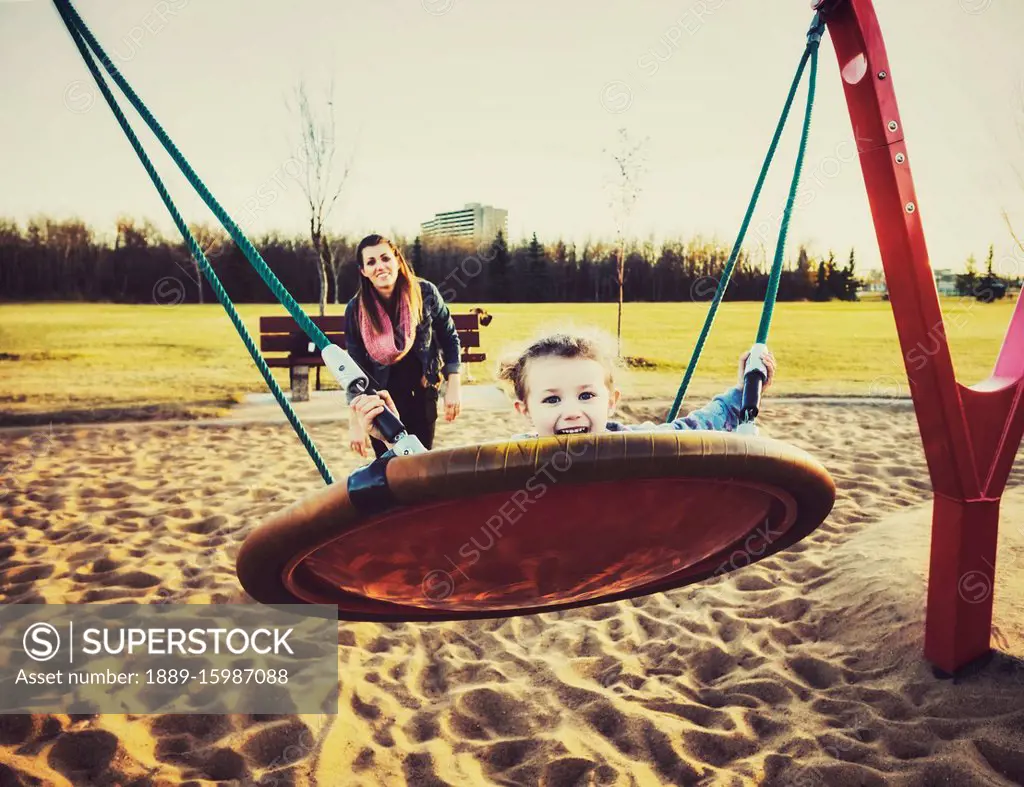 A young mom and her daughter playing on a saucer swing in a playground on a warm autumn evening; Edmonton, Alberta, Canada