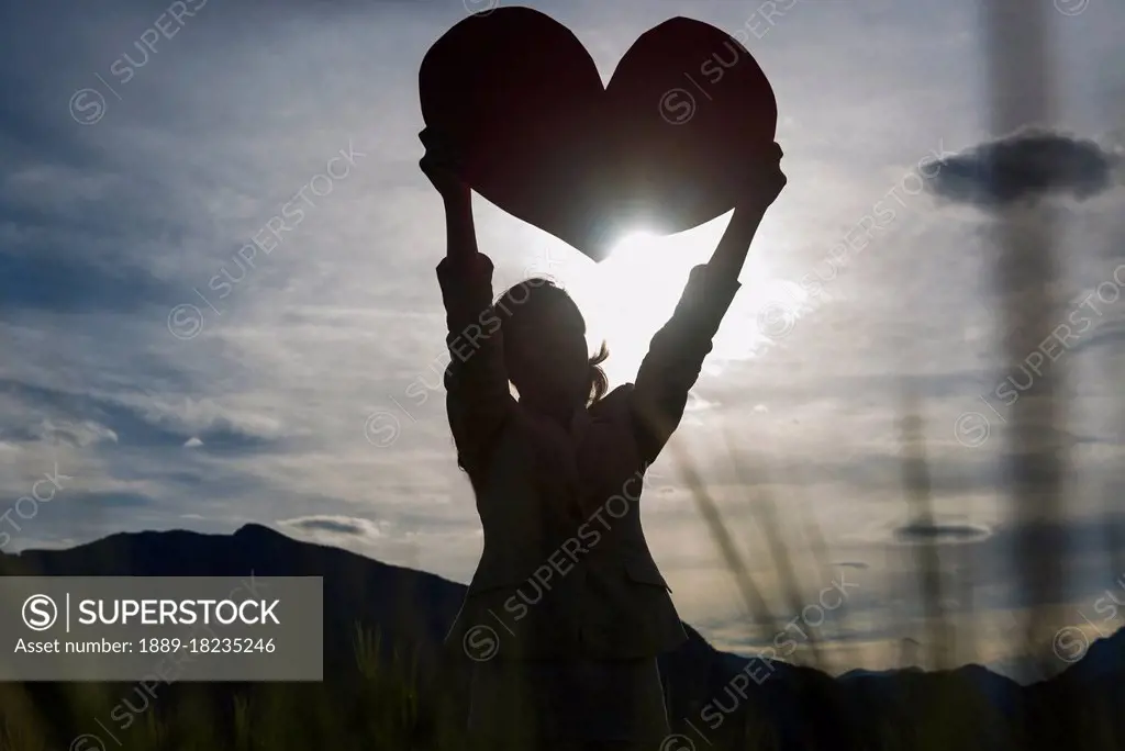 Silhouette of a woman holding up a large heart shape against the sunlight
