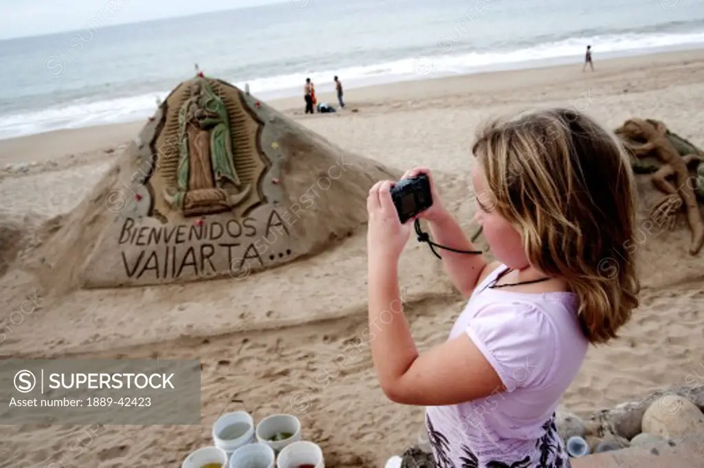 Malecon, Puerto Vallarta, Mexico; Young girl taking photo of sand sculpture