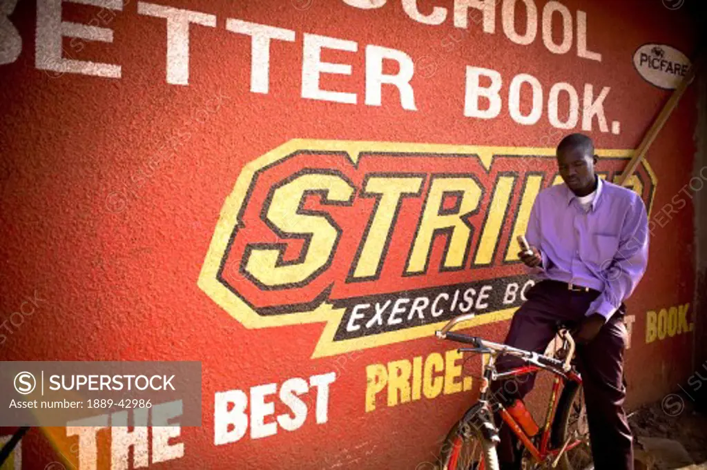 Gulu, Northern Uganda; Man sitting on bicycle beside sign, text messaging on mobile phone