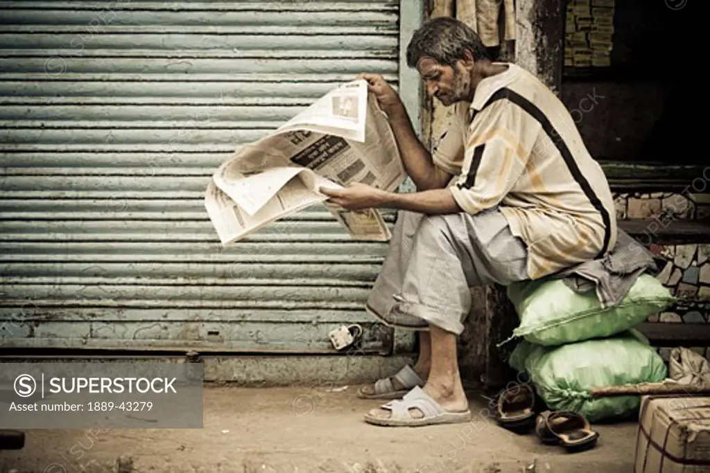 Delhi, India; Man sitting on sack reading newspaper