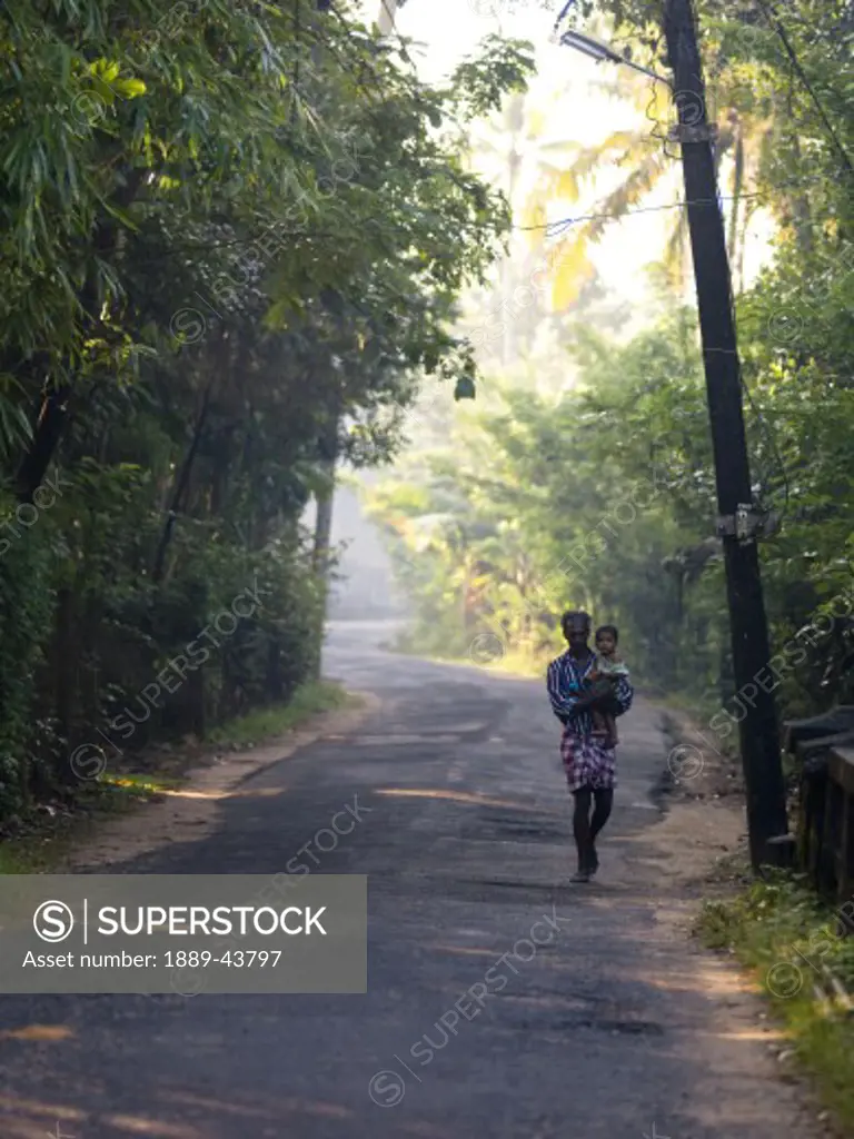 Kerala,India;Father carrying baby daughter down lush tree lined road