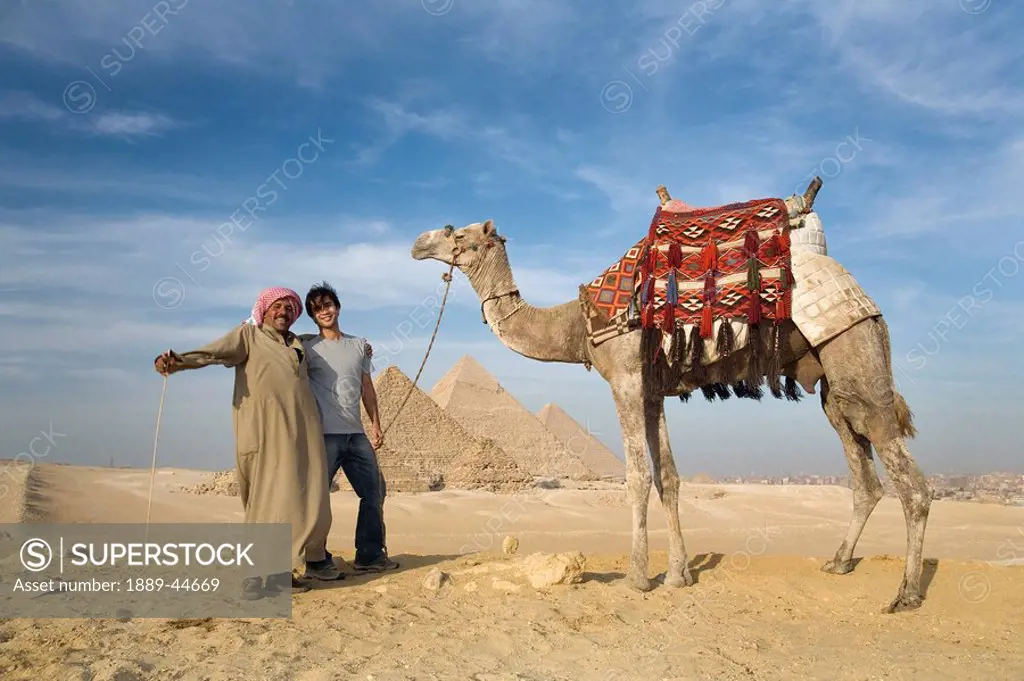 Two men in the desert with a camel with the Pyramids in the background