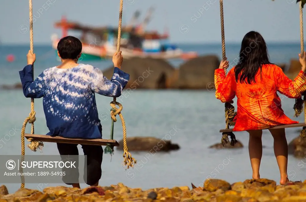 Thailand, Two People Sitting On Swings On The Coast Watching A Boat In The Water