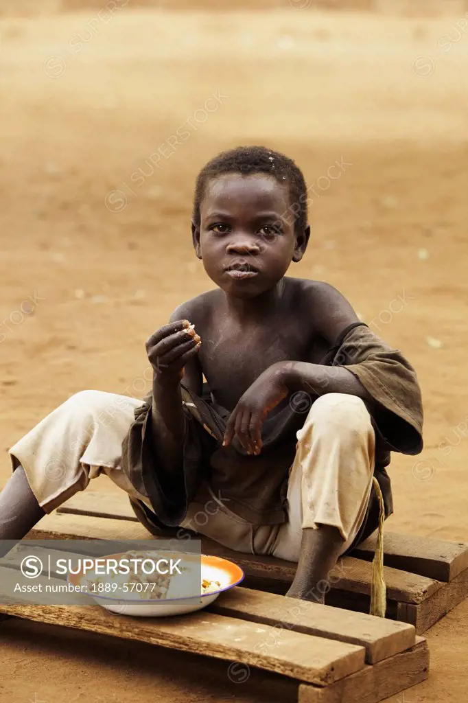 A Boy Eats A Meal Sitting On Boards On The Ground, Manica, Mozambique, Africa