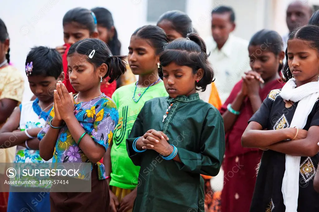girls standing in rows with eyes closed in prayer with men standing in the background, sathyamangalam, tamil nadu, india