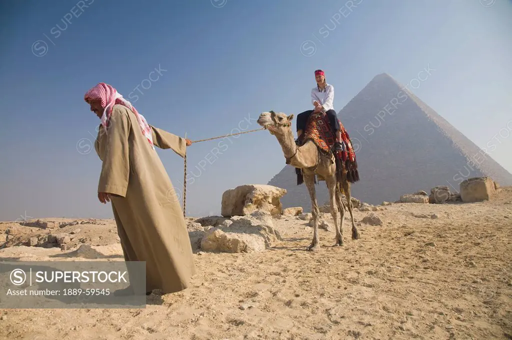 Young woman tourist riding a camel lead by a guide at the Pyramids of Giza, Egypt