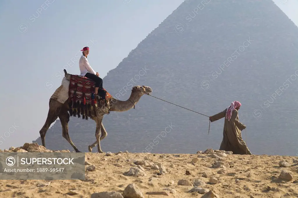Young woman tourist riding a camel lead by a guide at the Pyramids of Giza, Egypt