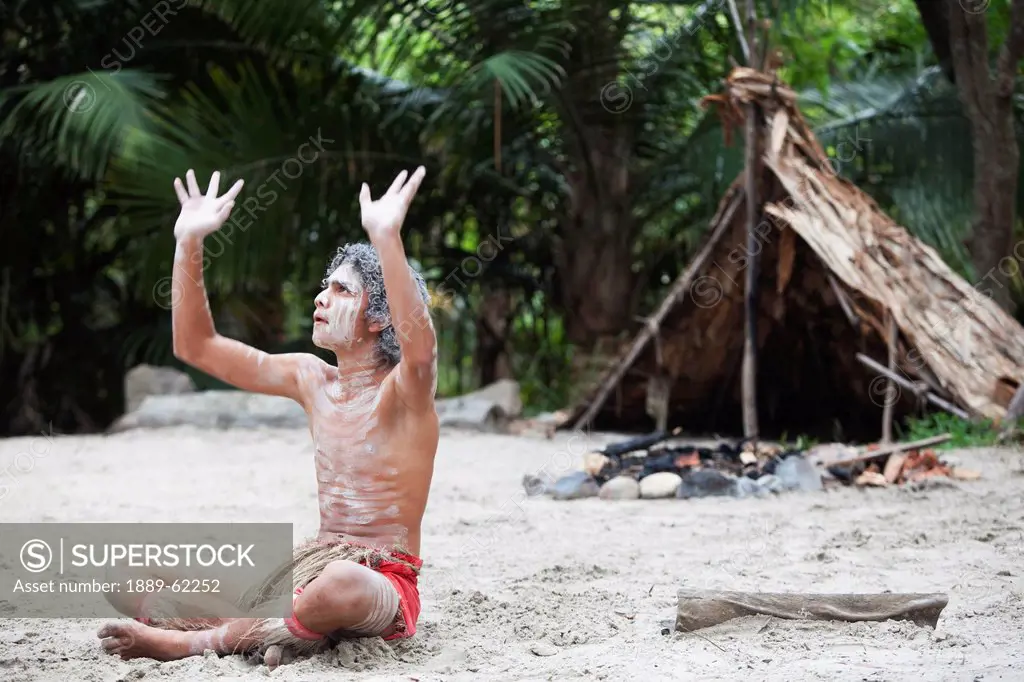 an indigenous man siting on the sand with hands raised to the sky, queensland, australia