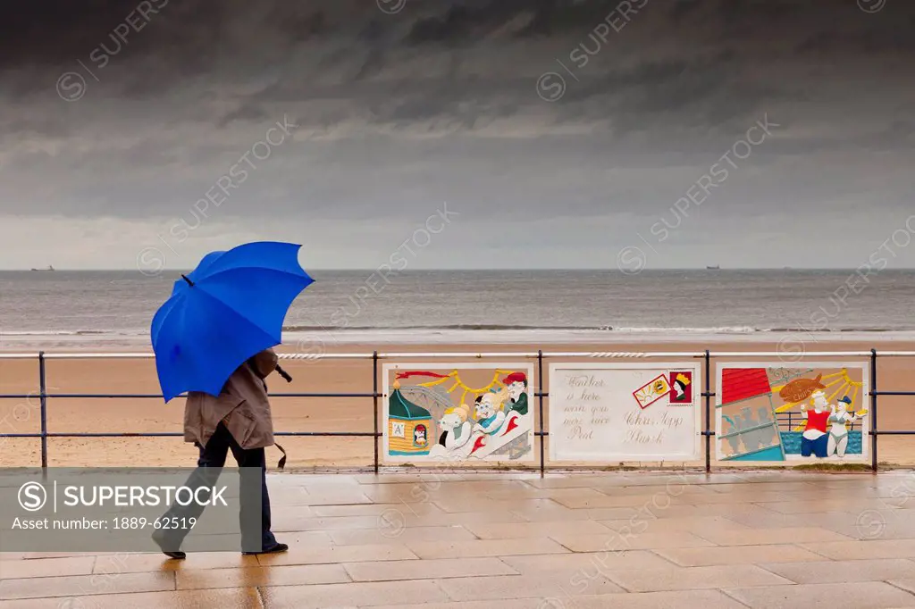 a person walks with an umbrella on a promenade along the water on a rainy day, redcar, teesside, england
