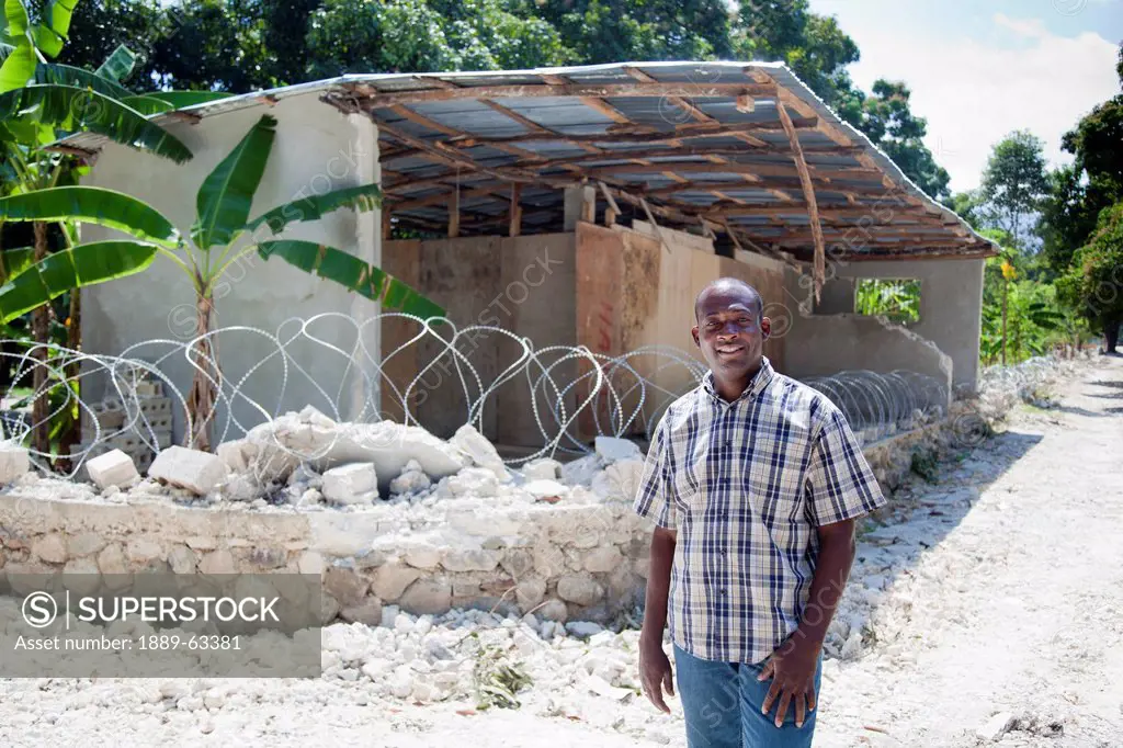 a man stands in front of ruins and a barbed wire sitting on top of what was a compound wall, grand goave, haiti