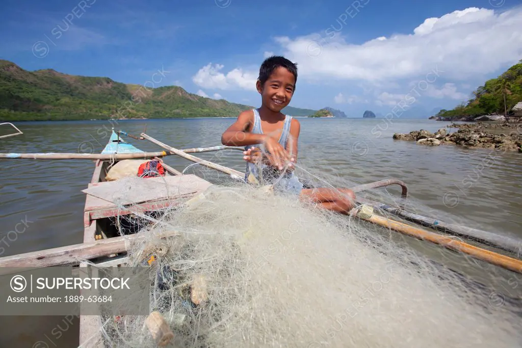 a young filipino boy untangles fish nets on his family´s wooden bangka boat in the tiny fishing village of vigan near snake island and el nido, vigan,...
