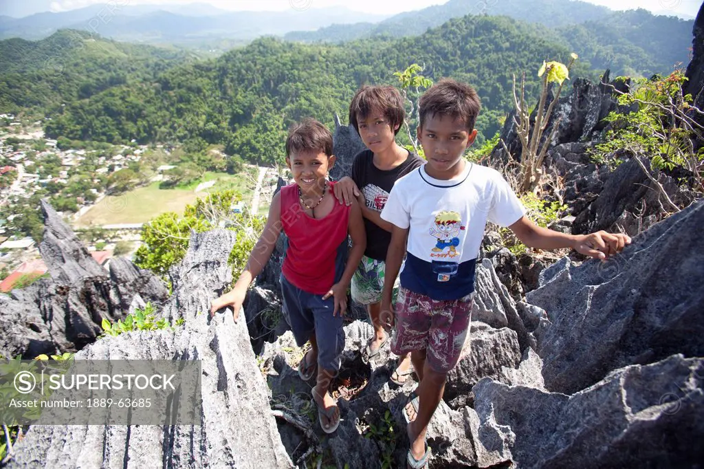 three young boys pose for a photograph as they stand high up on limestone spires overlooking the village and tourist destination of el nido, el nido, ...