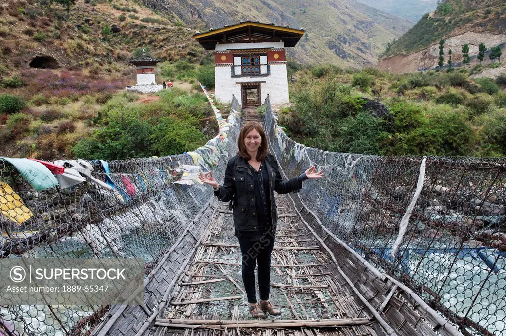 a woman stands on an iron suspension bridge lined with prayer flags, tamchhog lhakhang bhutan