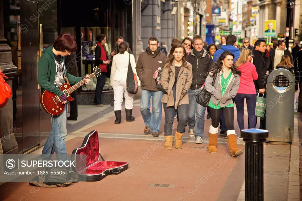 a busker playing a guitar on the sidewalk as pedestrians walk by on grafton street, dublin ireland